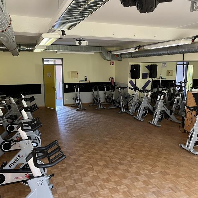 Fitnessraum mit mehreren Spinning-Bikes auf einem Holzfußboden und großen Spiegeln an den Wänden.