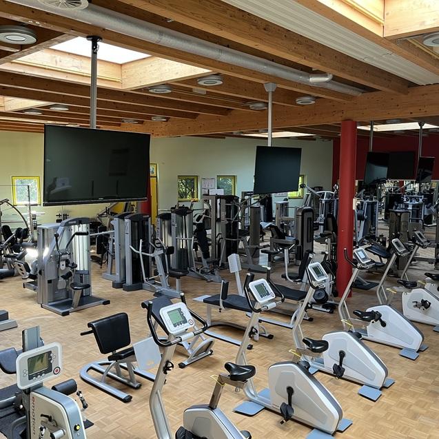 Innenansicht eines Fitnessstudios mit verschiedenen Trainingsgeräten und großen Fenstern, die Tageslicht hereinlassen.