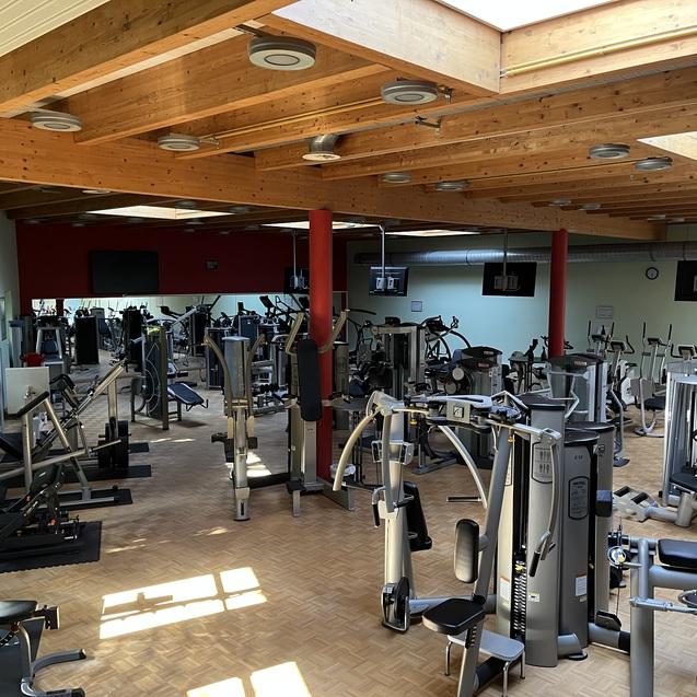 Weitblick auf einen gut ausgestatteten Fitnessraum mit verschiedenen Trainingsgeräten und Holzdecken.