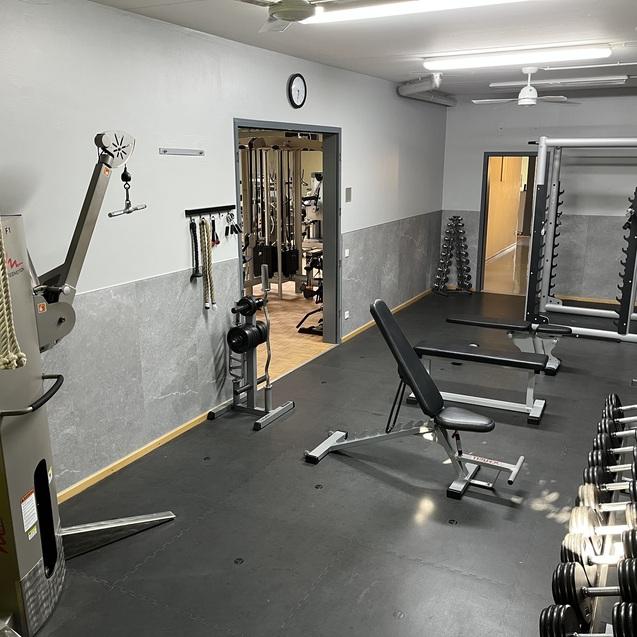 Fitnessraum mit verschiedenen Trainingsgeräten, einer Hantelbank und Gewichten auf dem Boden.