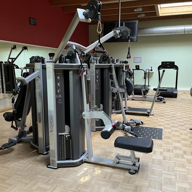 Fitnessstudio mit verschiedenen Krafttrainingsgeräten, Holzfußboden und Spiegel an der Wand.
