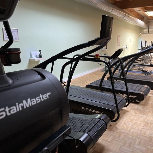 Blick auf mehrere Laufband-ähnliche Fitnessgeräte in einem hellen Trainingsraum mit Holzfußboden.
