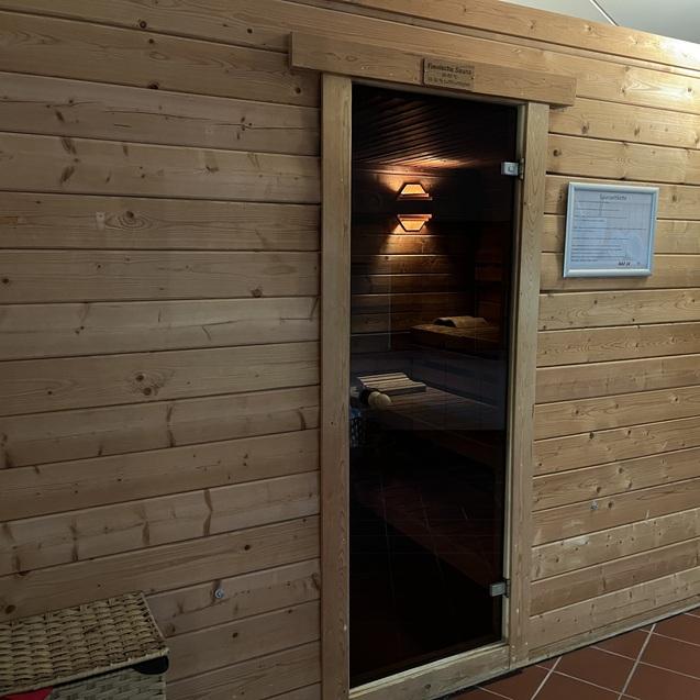 Holzsauna mit Glastür, betreten durch einen offenen Eingangsbereich, neben einem Korb auf gefliester Fläche.