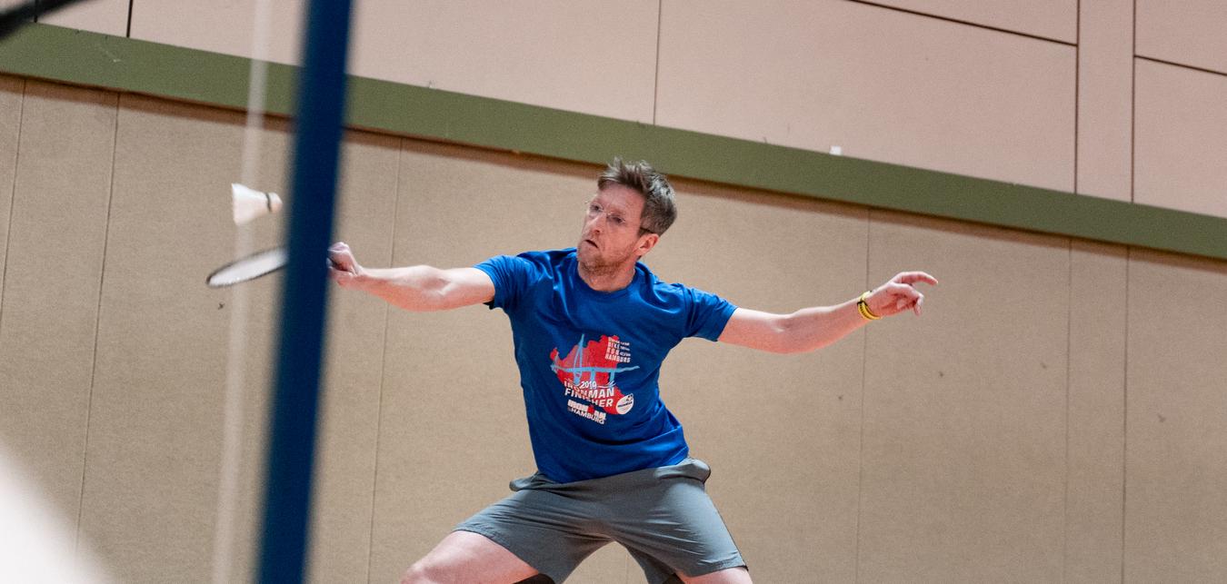 Mann in blauer T-Shirt und Shorts schl&auml;gt einen Federball in einer Sporthalle mit beige Wand.