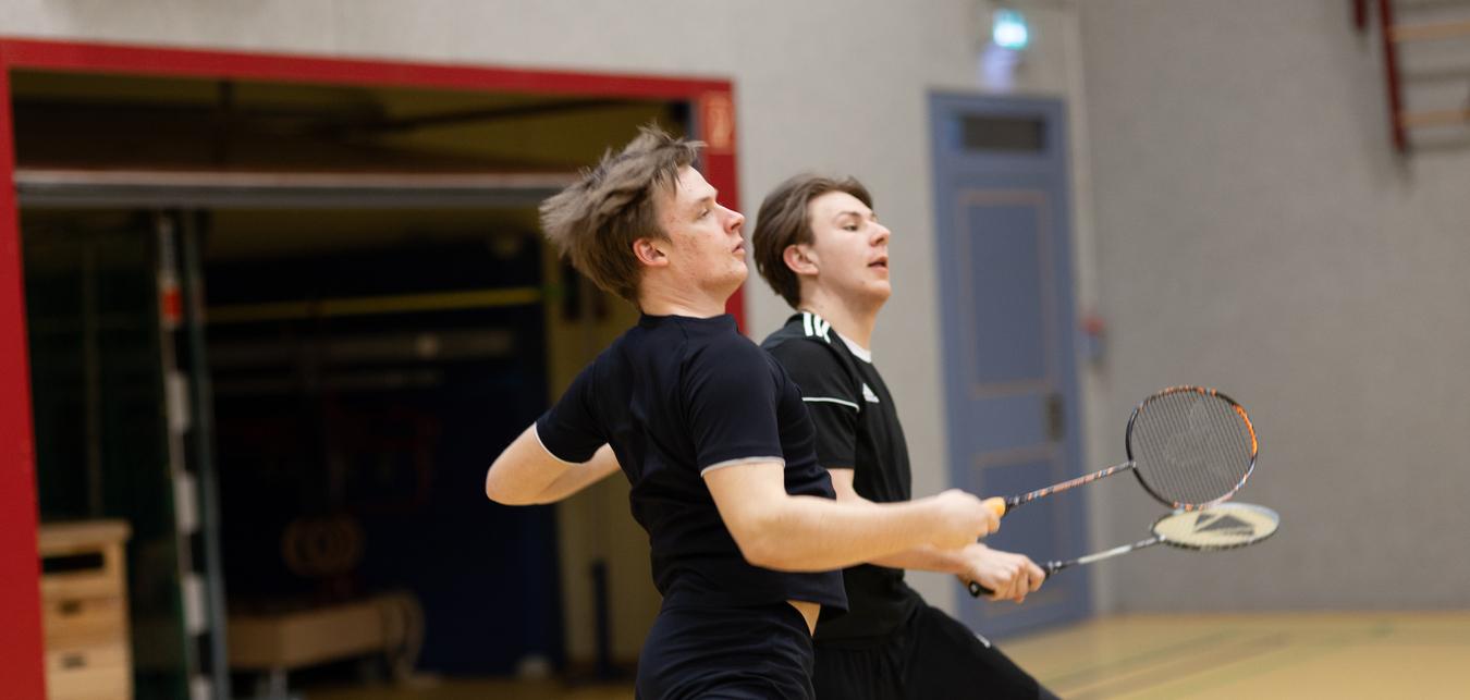 Zwei junge M&auml;nner spielen Badminton in einer Sporthalle, w&auml;hrend sie sich in Bewegung befinden.