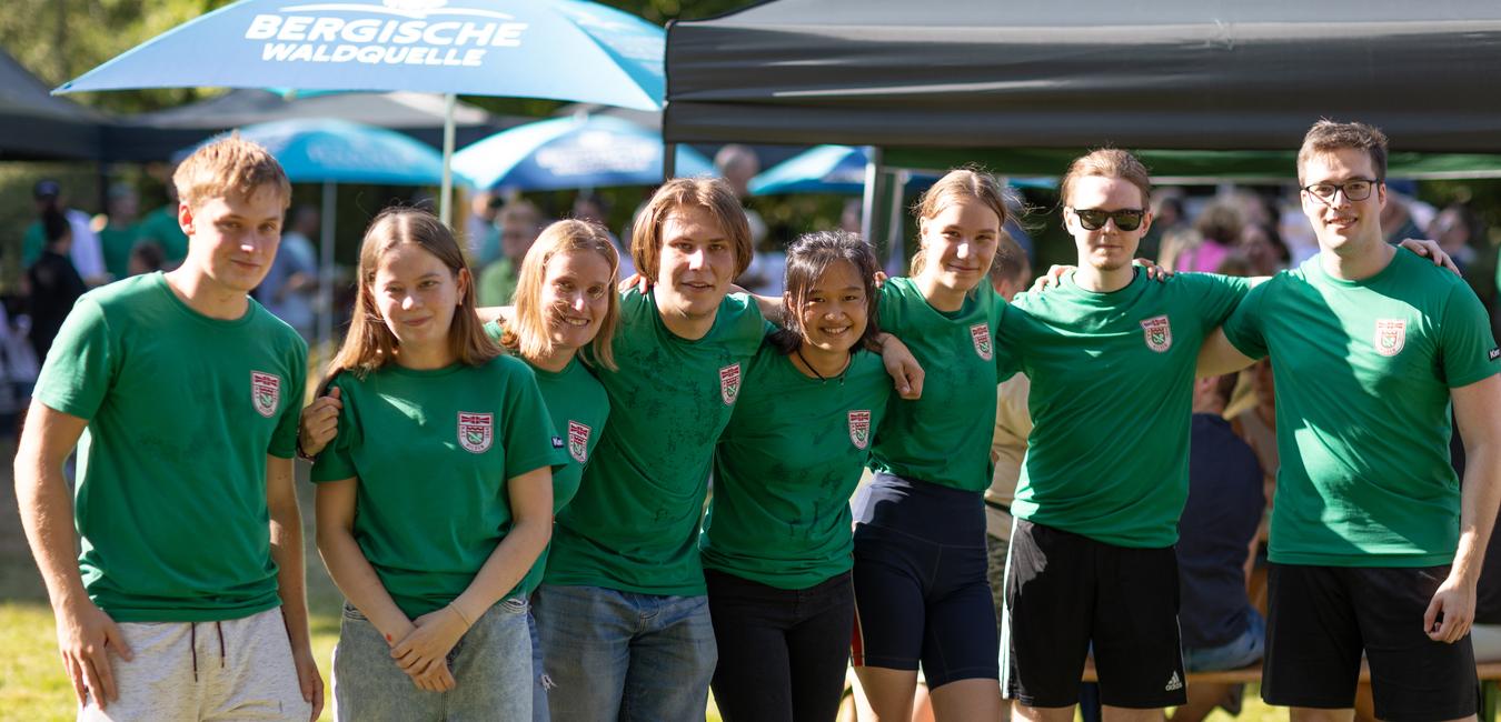 Gruppe von acht Personen in gr&uuml;nen T-Shirts, die in einer sonnigen Parklandschaft posieren.
