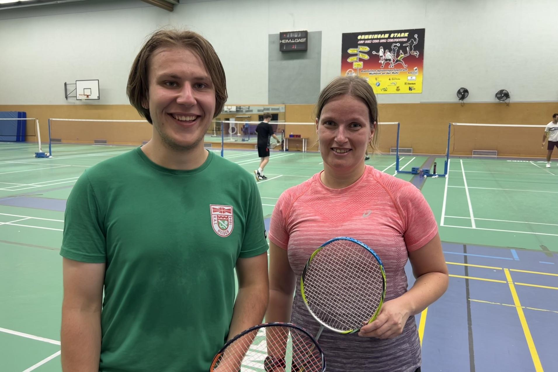 Zwei Badmintonspieler im Sportdress stehen lächelnd auf einem Badmintonfeld, jeder mit einem Schläger in der Hand.