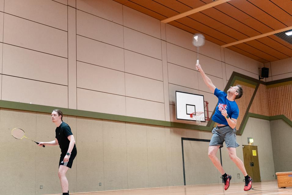 Zwei Personen spielen Badminton in einer Sporthalle; einer schl&auml;gt den Federball &uuml;ber das Netz.
