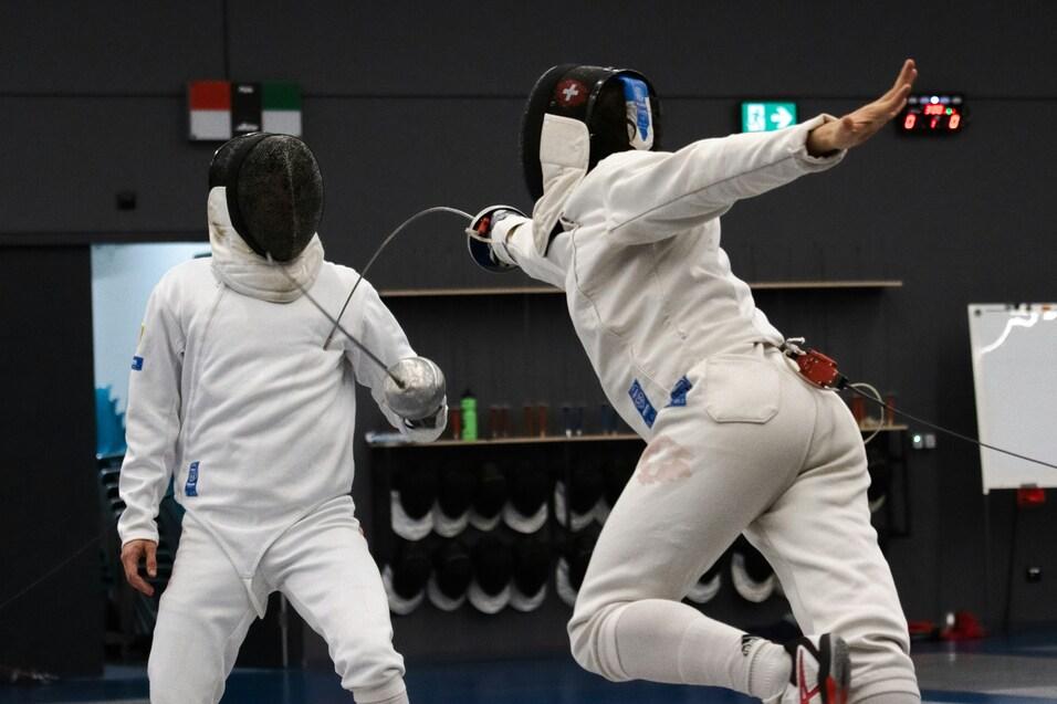 Zwei Fechter in vollständiger Ausrüstung kämpfen in einer Sporthalle, einer führt einen Schlag aus.