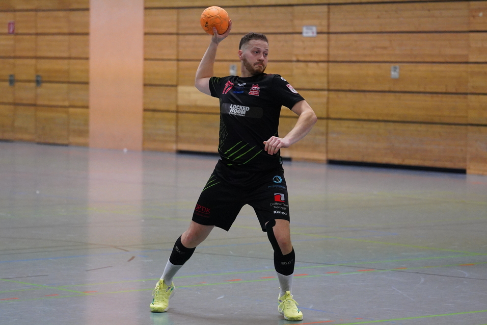 Spieler in schwarzem Trikot wirft einen Handball in einer Sporthalle, ein weiterer Spieler im Hintergrund in gelbem Trikot.