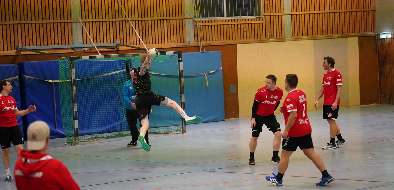 Spielszene in der Halle: Ein Spieler springt zum Wurf auf das Tor, umgeben von mitspielenden Handballern in roten Trikots.