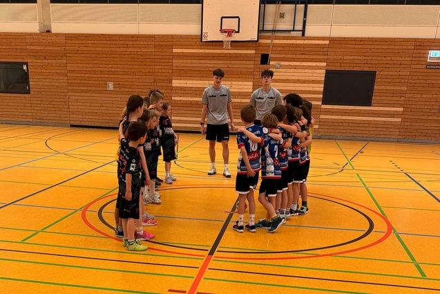 Zwei Gruppen von Kindern versammeln sich in einer Sporthalle zur Besprechung auf einem bunten Basketballfeld.