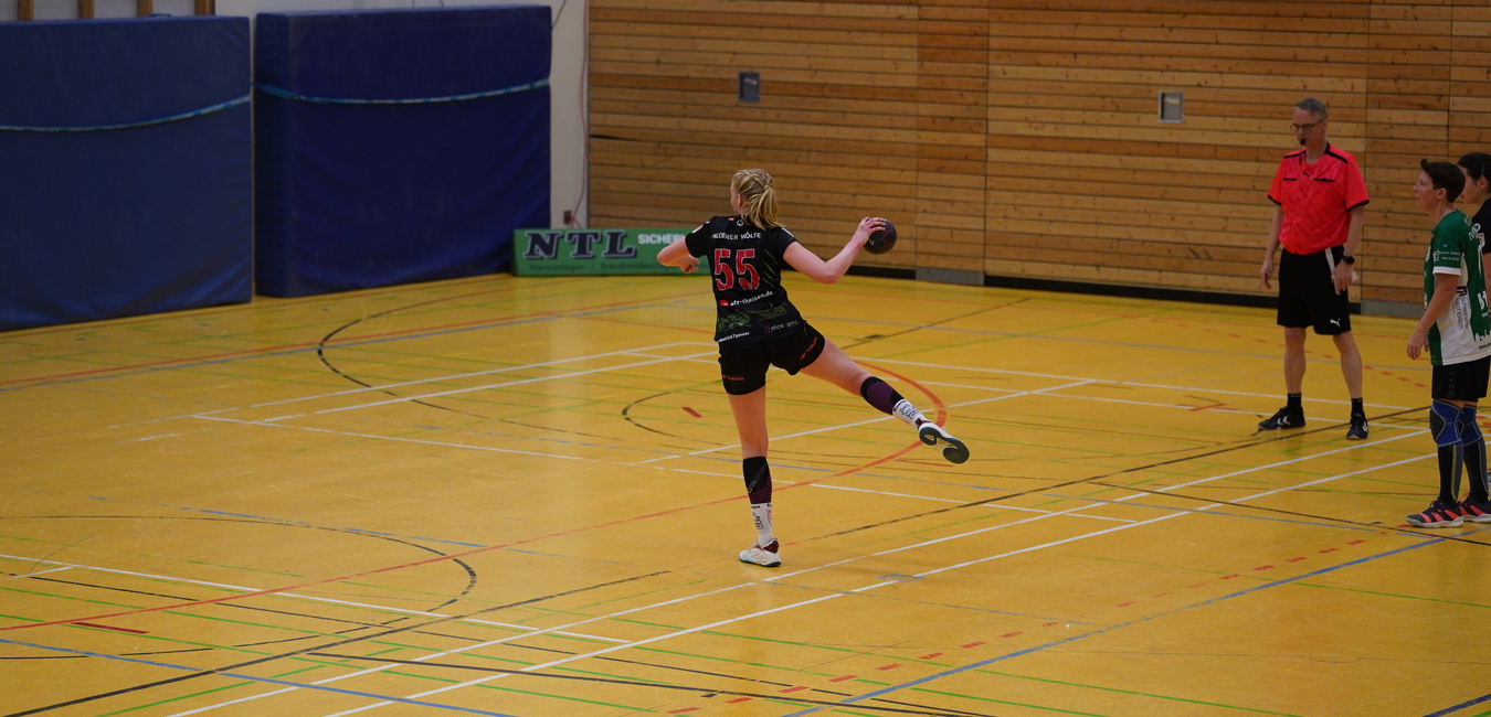 Handballspiel: Spielerin mit Trikotnummer 55 springt zum Wurf, w&auml;hrend Schiedsrichter und andere Spieler zuschauen.