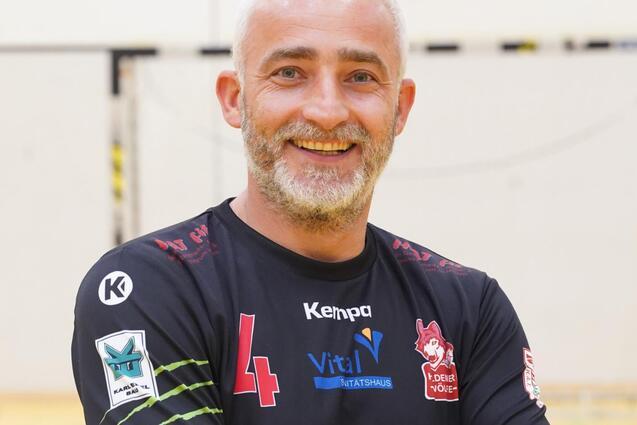 L&auml;chelnder Mann mit grauen Haaren, in schwarzem Sportshirt, steht vor einer Handball-Turnierfl&auml;che und verschr&auml;nkt die Arme.