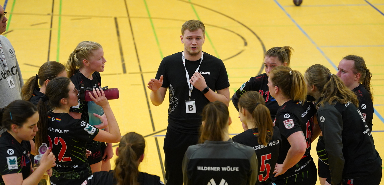 Trainer spricht mit einer Gruppe von weiblichen Handballspielern in einem Sporthallenraum.