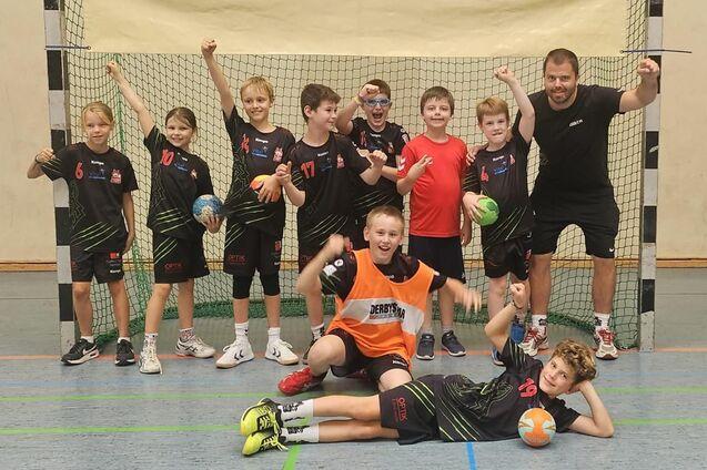 Gruppe von Kindern und einem Trainer in Sportkleidung vor einem Handballtor, einige halten B&auml;lle in den H&auml;nden.