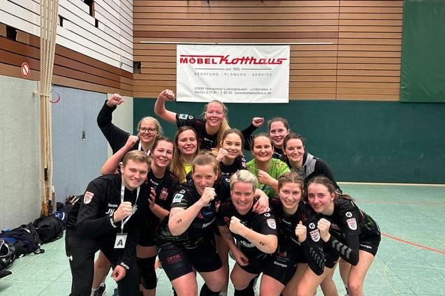Gruppensiegpose von Handballspielerinnen in einem Sportgymnasium, mit Punktestand und Werbebanner im Hintergrund.