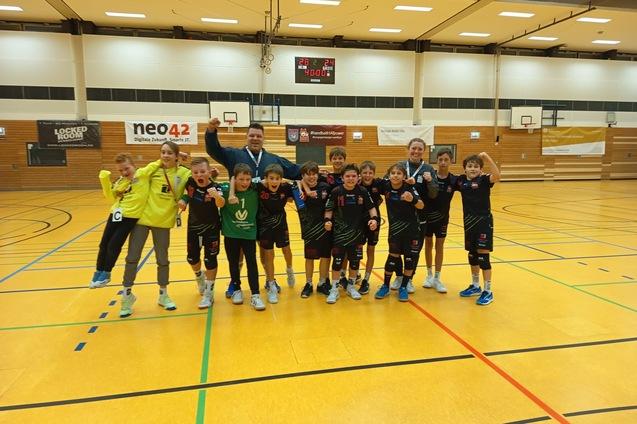 Gruppe von Kindern in Sportkleidung, l&auml;cheln und zeigen Medaillen in einer Turnhalle mit Basketballk&ouml;rben im Hintergrund.