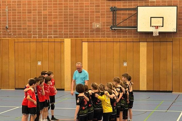 Zwei Gruppen von Kindern in Sportkleidung stehen sich in einer Sporthalle gegen&uuml;ber, ein Trainer gibt Anweisungen.