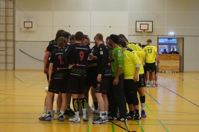 Eine Gruppe von Handballspielern in schwarzen Trikots steht um einen Trainer in gr&uuml;ner Kleidung auf dem Feld.