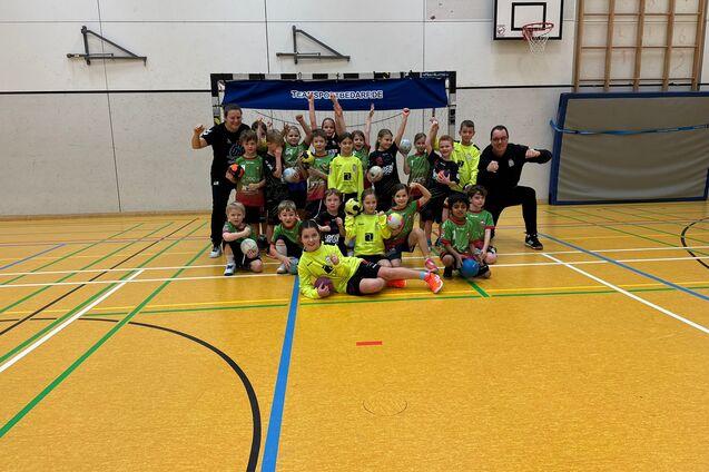 Gruppenfoto von Kindern in Sportkleidung mit Trainern in einer Sporthalle, alle posieren lachend mit B&auml;llen.