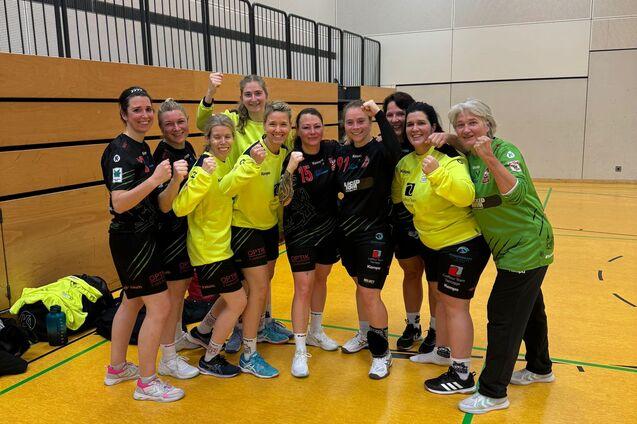 Gruppenfoto von zehn Handballspielerinnen in Sportkleidung, die im Trainingsraum posieren und zum Sieg zeigen.
