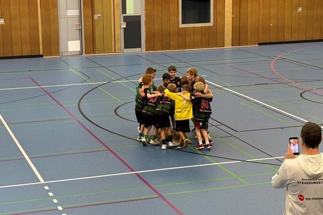 Gruppe von Kindern in Sportbekleidung, die sich in der Halle zum Team-Huddle umarmen. Ein Erwachsener macht ein Foto.