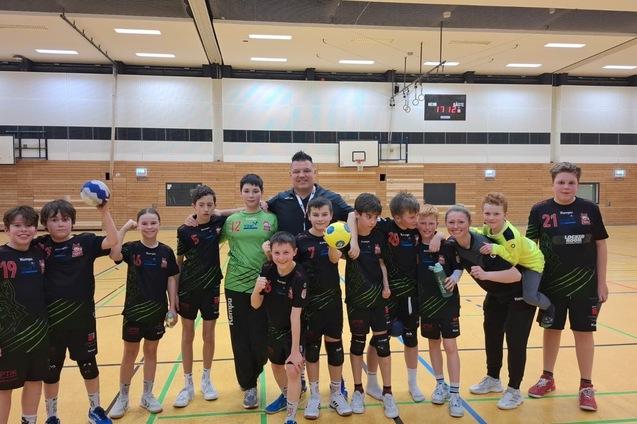 Gruppe von Jugendlichen in Sportkleidung, l&auml;chelnd auf einem Handballfeld mit B&auml;llen in der Hand.