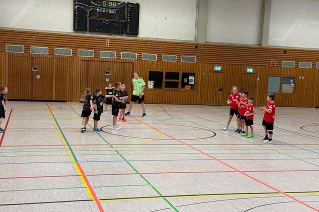 Zwei Gruppen von Kindern in Sportkleidung spielen Handball in einer hellen Halle mit markierten Linien auf dem Boden.
