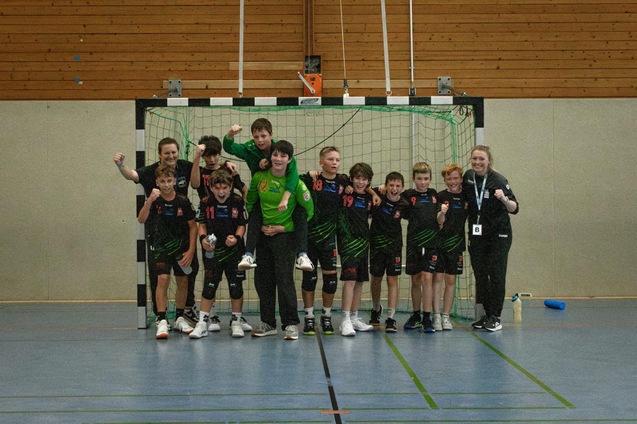 Gruppe von Handballspielern und Trainern posiert fr&ouml;hlich vor einem Tor in einer Sporthalle.