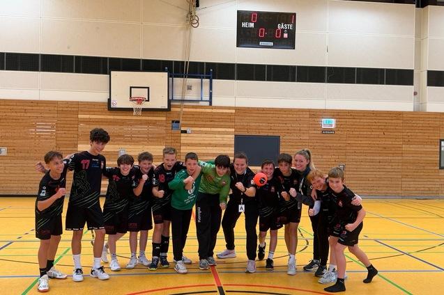 Gruppe von Handballspielern und Trainerin posieren l&auml;chelnd in sportlicher Kleidung auf dem Spielfeld einer Sporthalle.