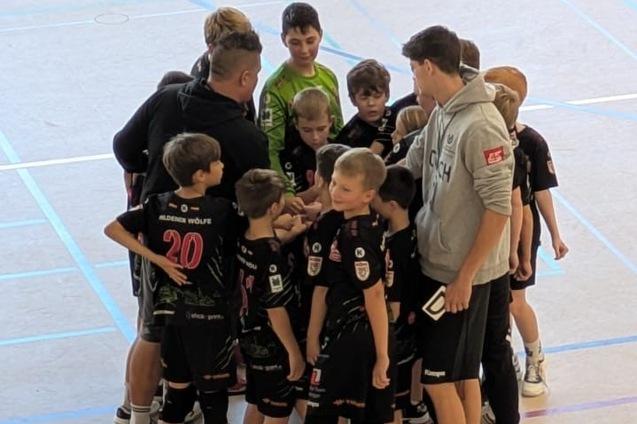 Gruppe von Jungen in Sportkleidung um einen Trainer versammelt, in einer Halle mit bunten Linien auf dem Boden.