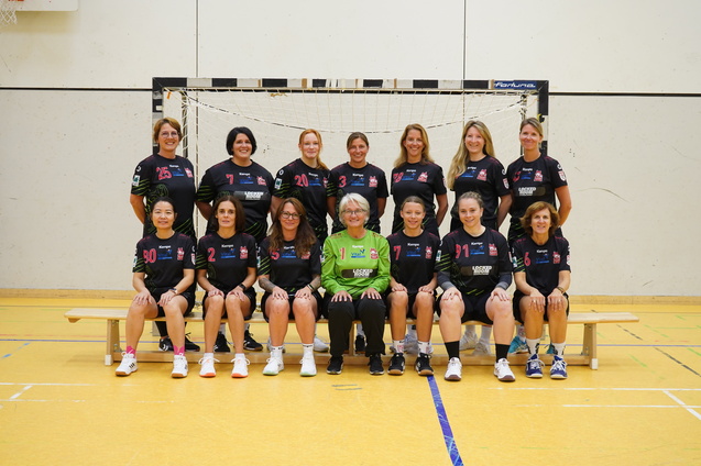 Gruppenfoto eines Handballteams mit 14 Spielerinnen und einer Trainerin in Sportkleidung vor einem Handballtor in einer Halle.