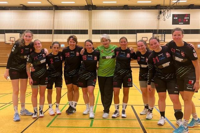 Gruppenshot einer Handballmannschaft in schwarz-roten Trikots, l&auml;chelnd auf einem Sportplatz in einer Halle.