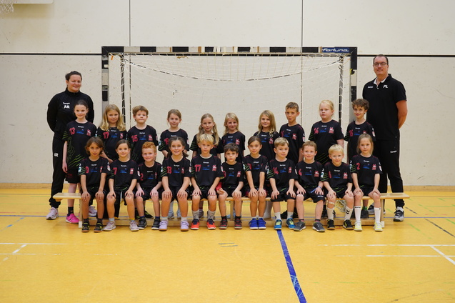 Gruppenbild einer Jugendmannschaft mit Trainer, alle in schwarzen Trikots vor einem Handballtor in einer Sporthalle.