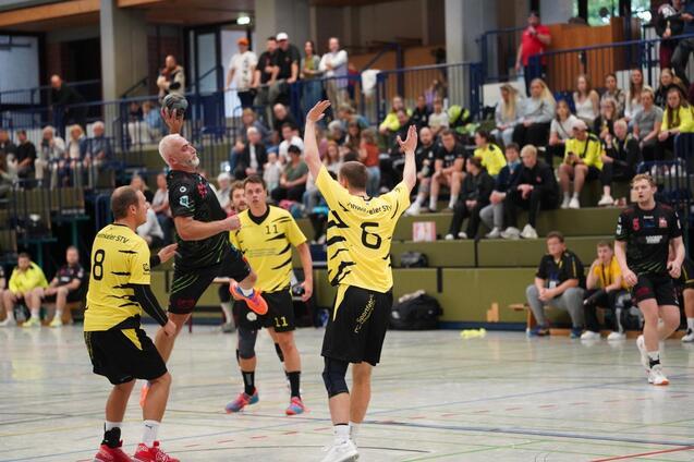Ein Handballspieler springt, um zu werfen, w&auml;hrend zwei verteidigende Spieler ihn anvisieren. Zuschauer im Hintergrund.