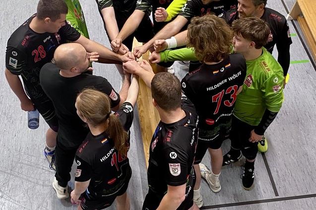 Gruppe von Handballspielern in schwarzen Trikots macht einen Teamkreis um eine Holzbank in einer Sporthalle.