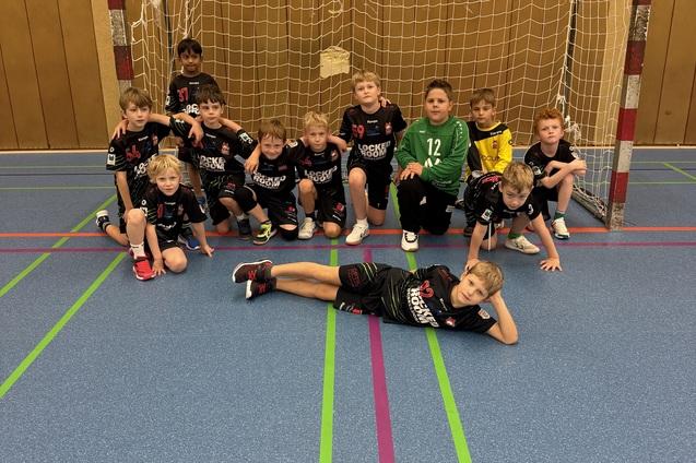 Gruppe von zw&ouml;lf Kindern in Sportkleidung, posierend vor einem Handballtor in einer Halle.