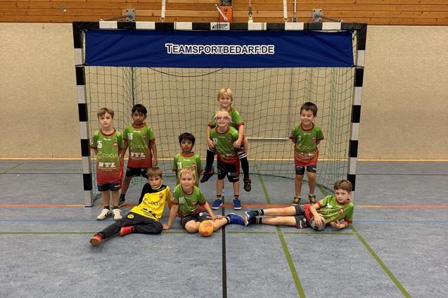 Gruppe von Kindern in Sportkleidung vor einem Handballtor in einer Turnhalle, einige sitzen, andere stehen.