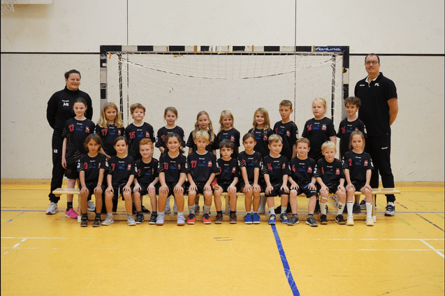 Gruppenfoto von Kindern in Sporttrikots, die vor einem Handballtor auf einer Sporthalle stehen.