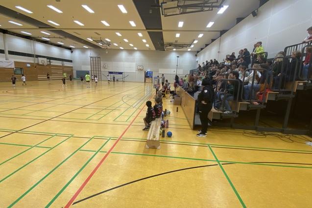 Sporthalle mit Kindern, die spielen und Zuschauern auf Trib&uuml;nen, Holzbank und Markierungen auf dem Boden.