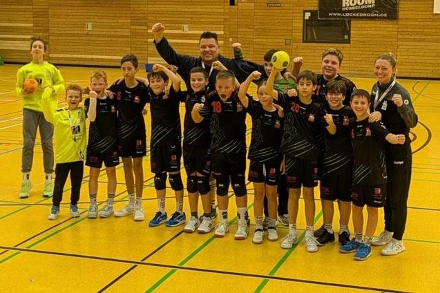 Gruppe jubelnder Kinder, Trainer und Betreuer in Sportkleidung auf einem Handballfeld.