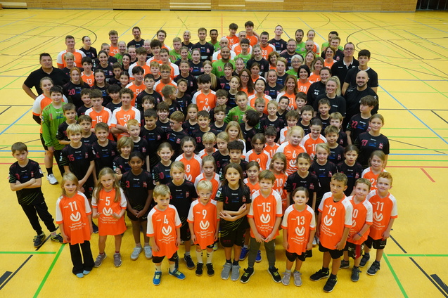 Gruppenfoto von Kindern und Erwachsenen in Sporttrikots auf einem gelben Hallenboden, l&auml;chelnd und aufgestellt.