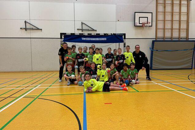 Gruppe von Kindern in Sportkleidung, die in einer Turnhalle vor einem Fu&szlig;balltor posieren. Trainer stehen hinter ihnen.