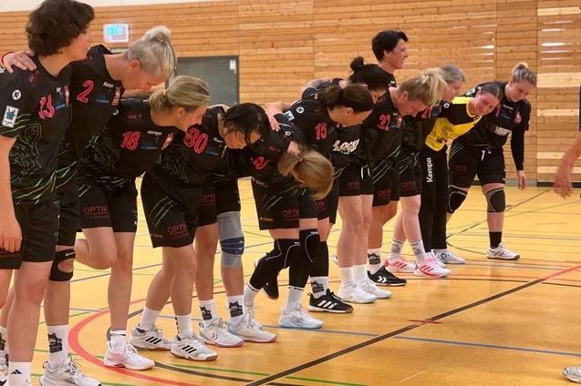 Frauen-Handballteam in Spielkleidung verbeugt sich am Spielfeldrand. Holzboden und Sportger&auml;te sind im Hintergrund sichtbar.