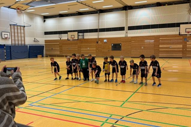 Gruppe von Kindern in Sportkleidung steht auf einer Turnhalle mit Linien und Basketballschultern im Hintergrund.