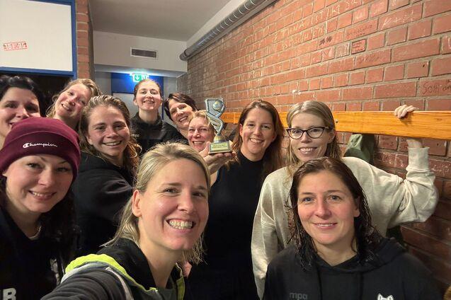 Gruppe von zehn Frauen posiert l&auml;chelnd f&uuml;r ein Selfie in einer sportlichen Umgebung mit einer Troph&auml;e im Hintergrund.