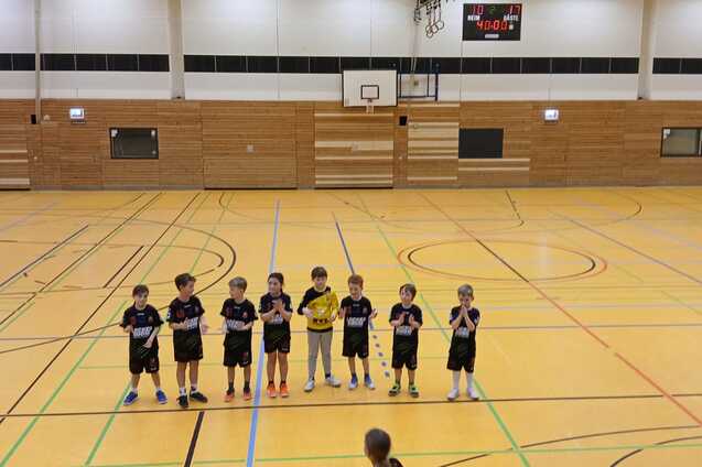 Gruppe von sieben Kindern in Sportkleidung steht auf einem Holzfu&szlig;boden in einer Sporthalle.