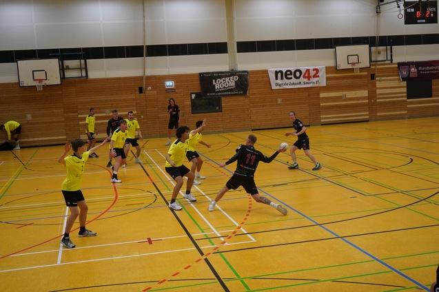 Spielszene aus einer Sporthalle: Handballspieler in gelben Trikots in Aktion, ein Spieler in Schwarz wirft den Ball.