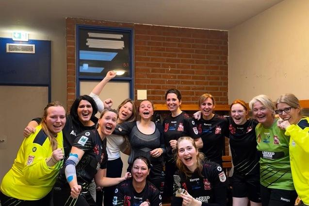 Gruppe von 13 jubelnden Frauen in Sportkleidung stehen freudig in einem Umkleideraum.
